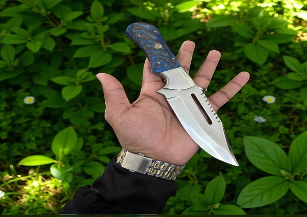 The Frostfang knife by Arham Cutlery featuring a blue pine cone resin handle and a D2 steel blade, displayed in hand against a green foliage background.