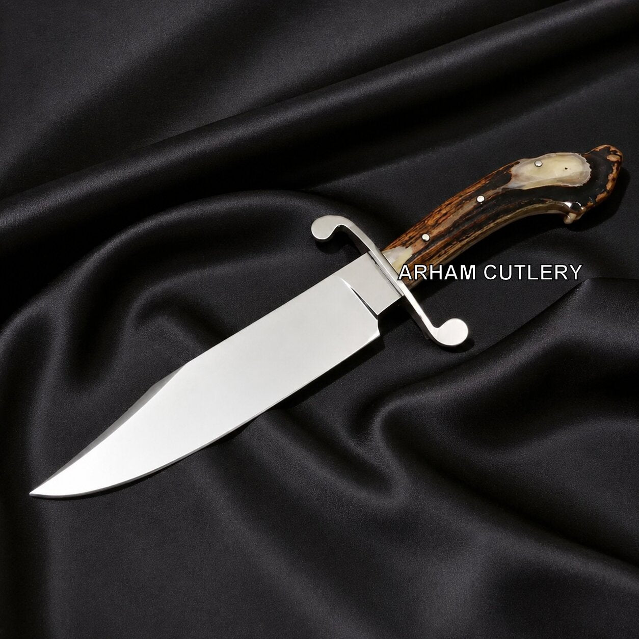 A custom fixed-blade knife with a polished silver blade, a curved silver guard, and a brown antler handle with a cream inlay, displayed on a draped black satin fabric background. The text 'ARHAM CUTLERY' is overlaid on the handle.