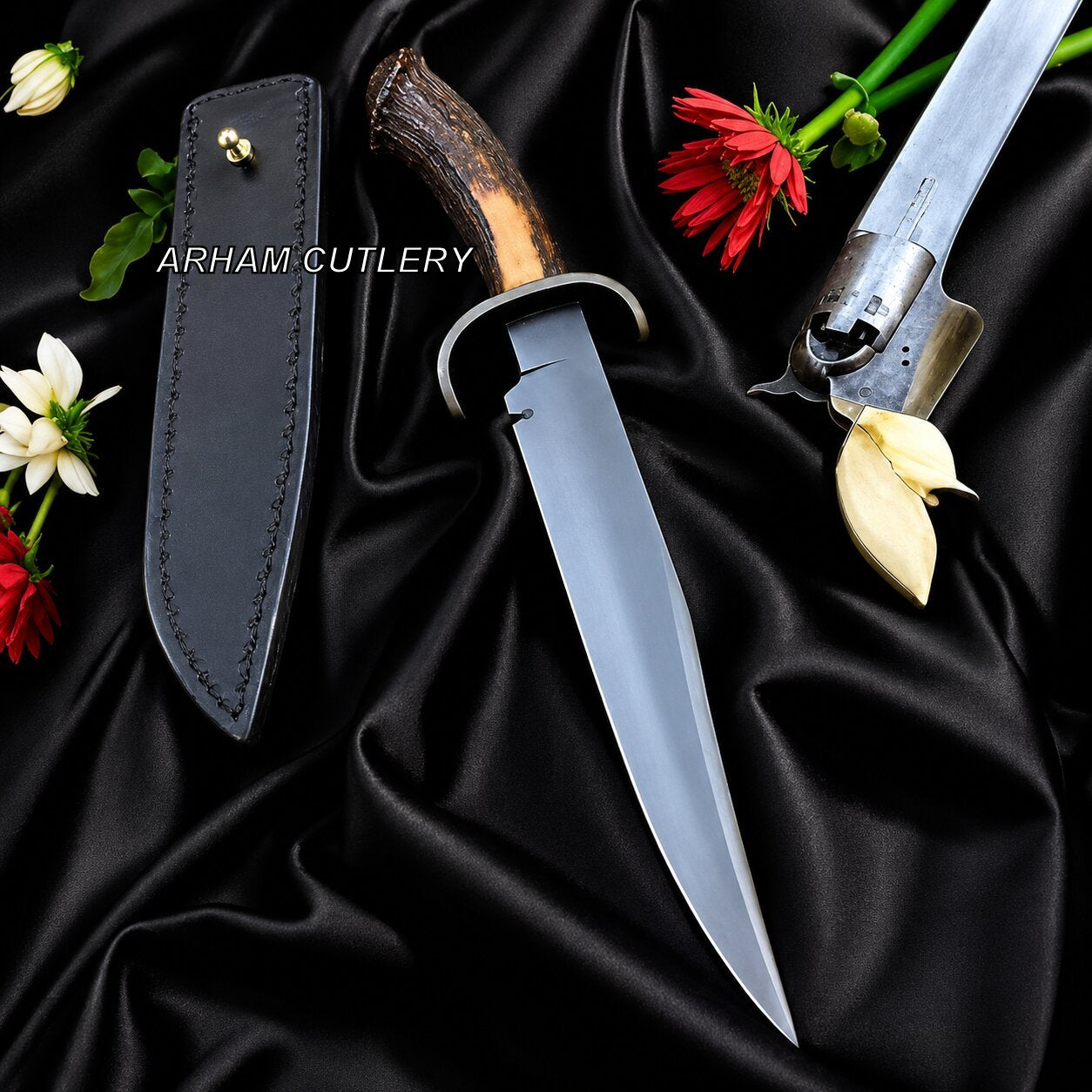 A fixed-blade hunting knife with a dark brown antler-style handle and a matte gray blade, displayed next to its black leather sheath on a draped black satin fabric. Red and white flowers are arranged as props, and the brand name 'ARHAM CUTLERY' is printed on the sheath.