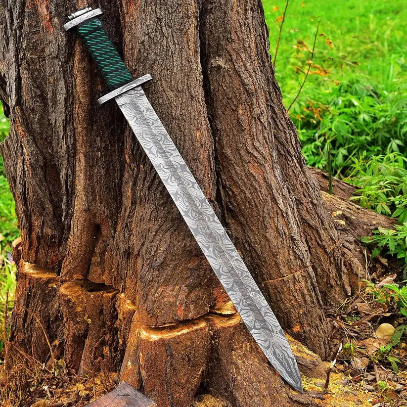 A decorative sword with a patterned steel blade and a green wrapped handle, leaning against a large tree trunk outdoors.