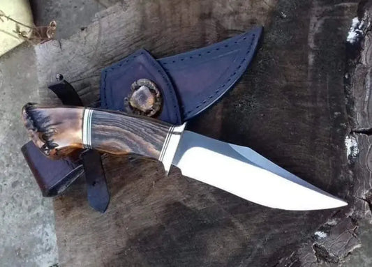 A fixed-blade knife with a polished silver blade and a carved brown handle, shown resting on a dark wooden surface next to a dark leather sheath.
