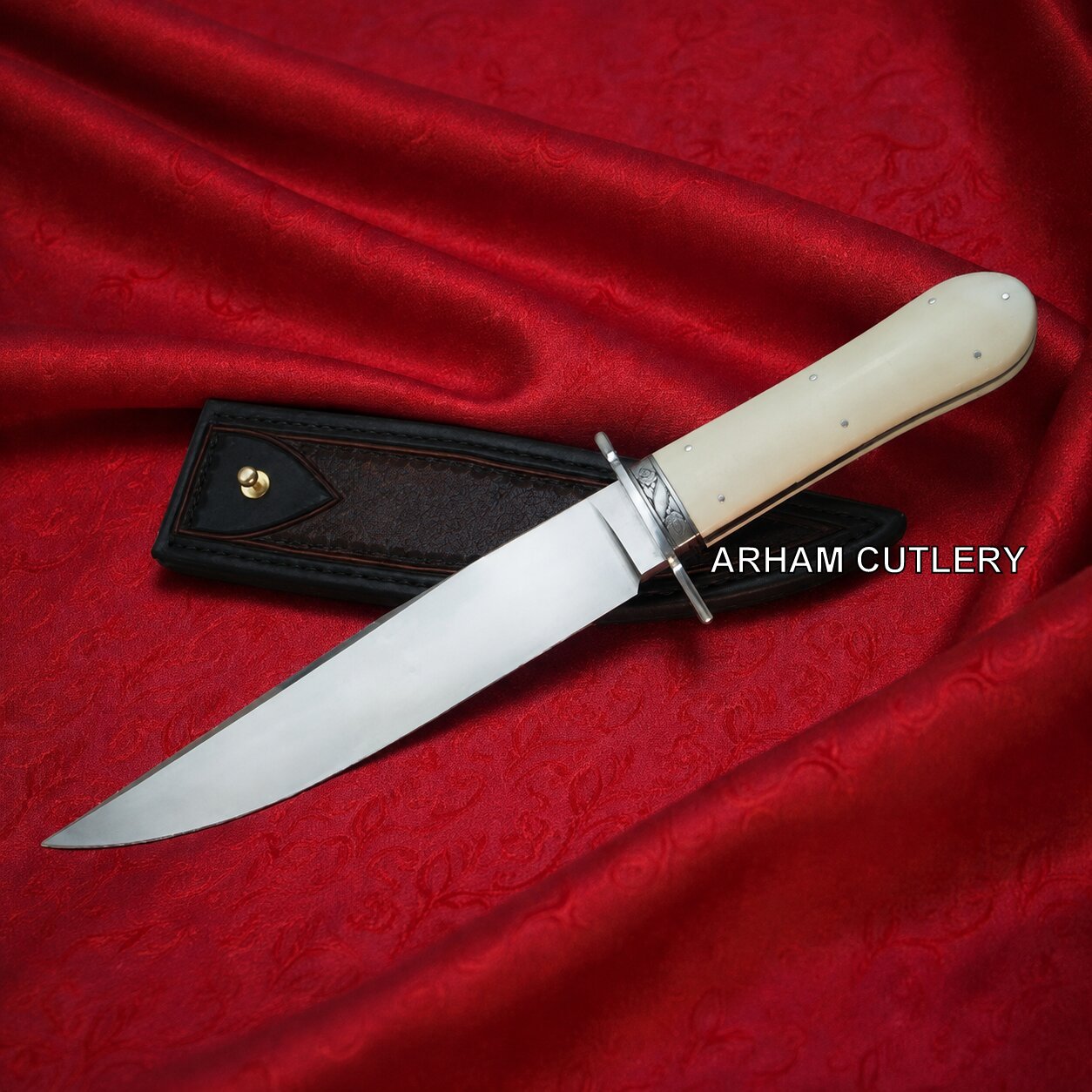 A fixed-blade knife with a polished silver blade, cream-colored bone handle, and silver guard, displayed diagonally on a textured red satin fabric next to its black leather sheath. The brand name 'ARHAM CUTLERY' is printed in white text over the handle area.