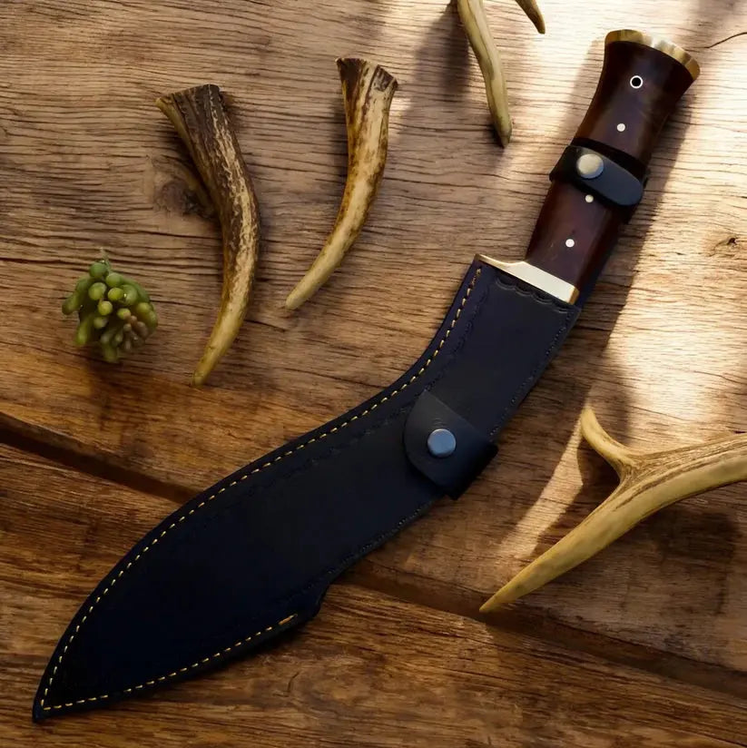 A fixed-blade knife with a dark brown wooden handle and black leather sheath, arranged diagonally on a rustic wooden surface with antlers and a small green sprig as props.