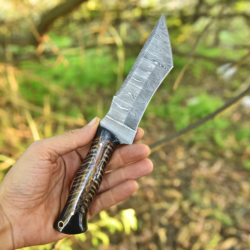 A hand holding a fixed-blade knife with a patterned Damascus-style steel blade and a dark brown and black striped handle, set against a blurred outdoor forest background.