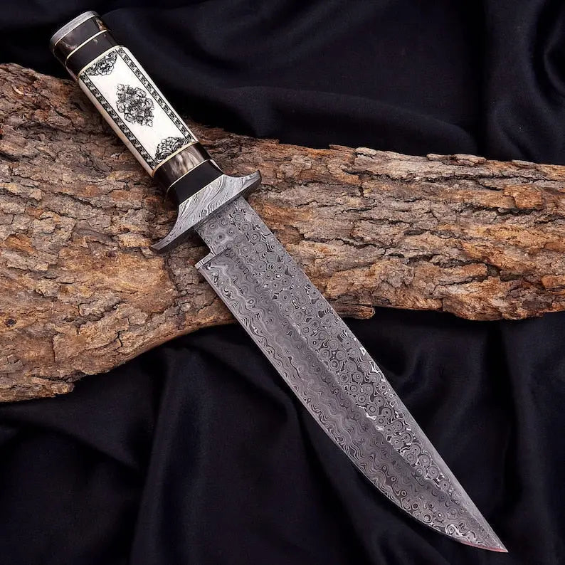 A custom handmade knife with a Damascus steel blade and an ornate handle, resting on a piece of tree bark over a black fabric background.