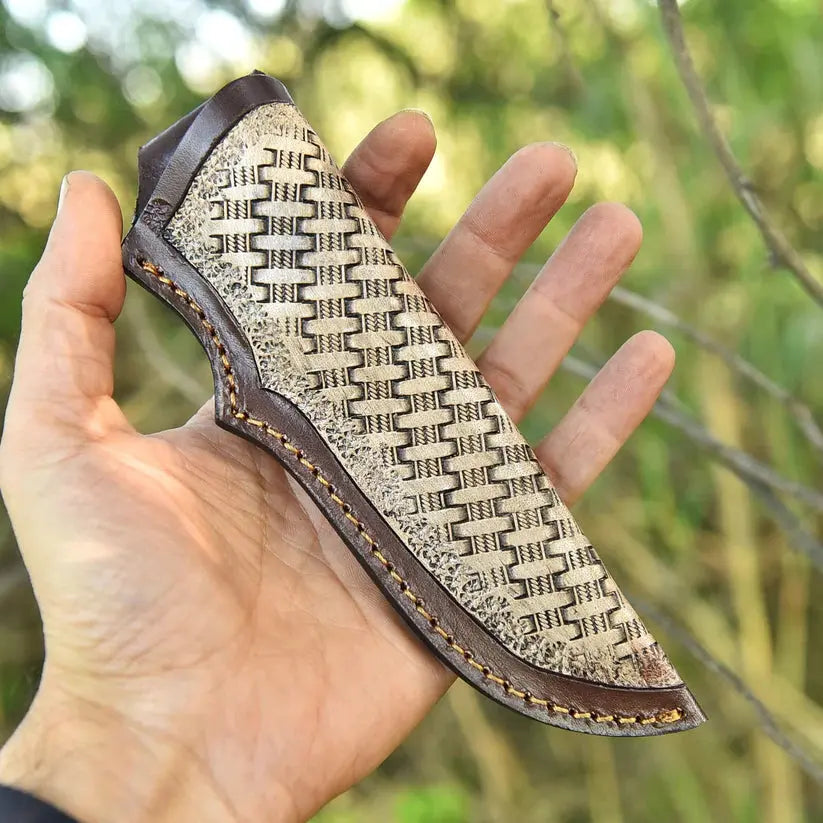 A hand holding a small knife sheath outdoors, with a blurred green and brown natural background. The sheath has a woven beige and dark brown pattern with dark brown leather trim and visible stitching.