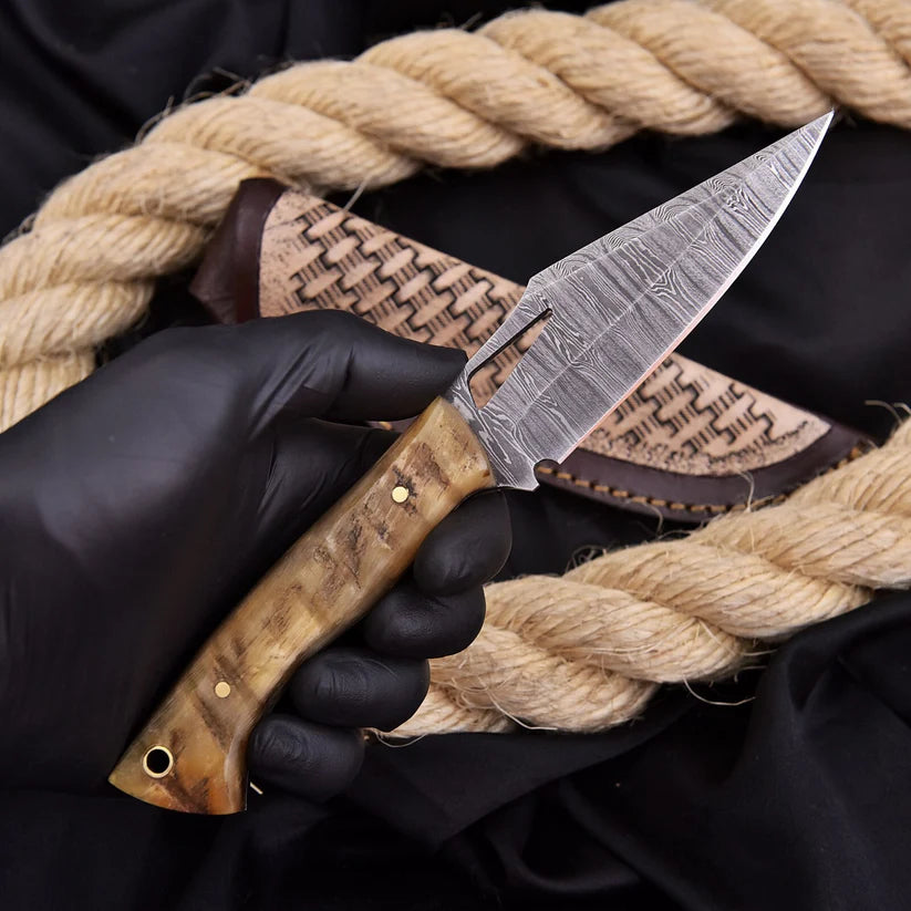 A hand wearing a black glove holds a fixed-blade knife with a patterned steel blade and a carved wooden handle, positioned in front of a woven sheath and thick natural rope on a black fabric background.