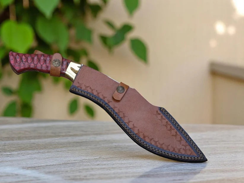 A fixed-blade knife with a textured reddish-brown handle and a brown leather sheath, resting on a light wooden surface with a blurred background of green leaves and a beige wall.