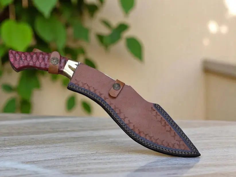 A fixed-blade knife with a textured reddish-brown handle and a brown leather sheath, resting on a light wooden surface with a blurred background of green leaves and a beige wall.