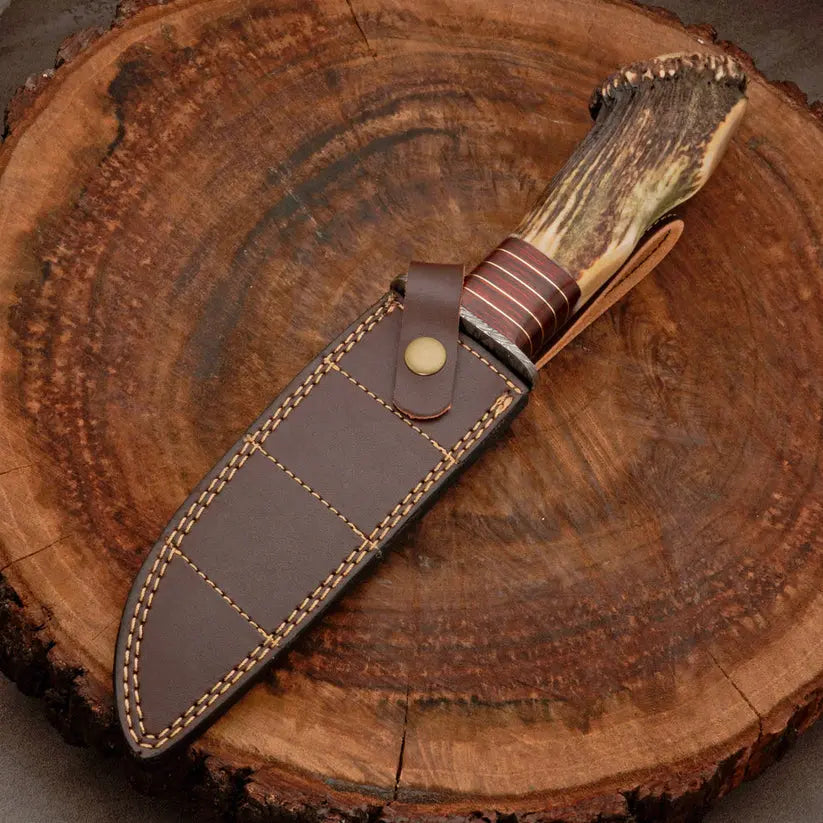 A fixed-blade knife with a natural antler-style handle and a dark brown leather sheath, displayed diagonally on a rustic wood log slice background.