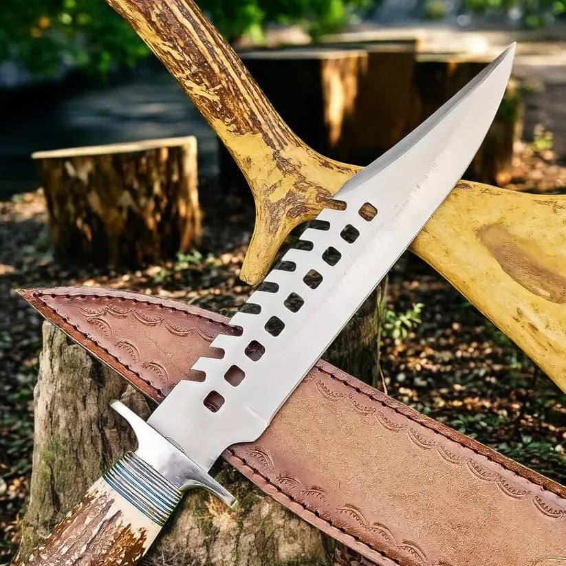 A fixed-blade knife with a silver blade featuring a distinctive square-hole cutout pattern along the spine, resting on a brown leather sheath. The knife is set against a rustic outdoor background with tree stumps and a large antlered axe handle.