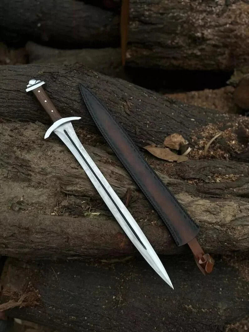 A double-edged dagger with a silver blade and brown handle lies next to its dark brown leather sheath on a rustic log outdoors.