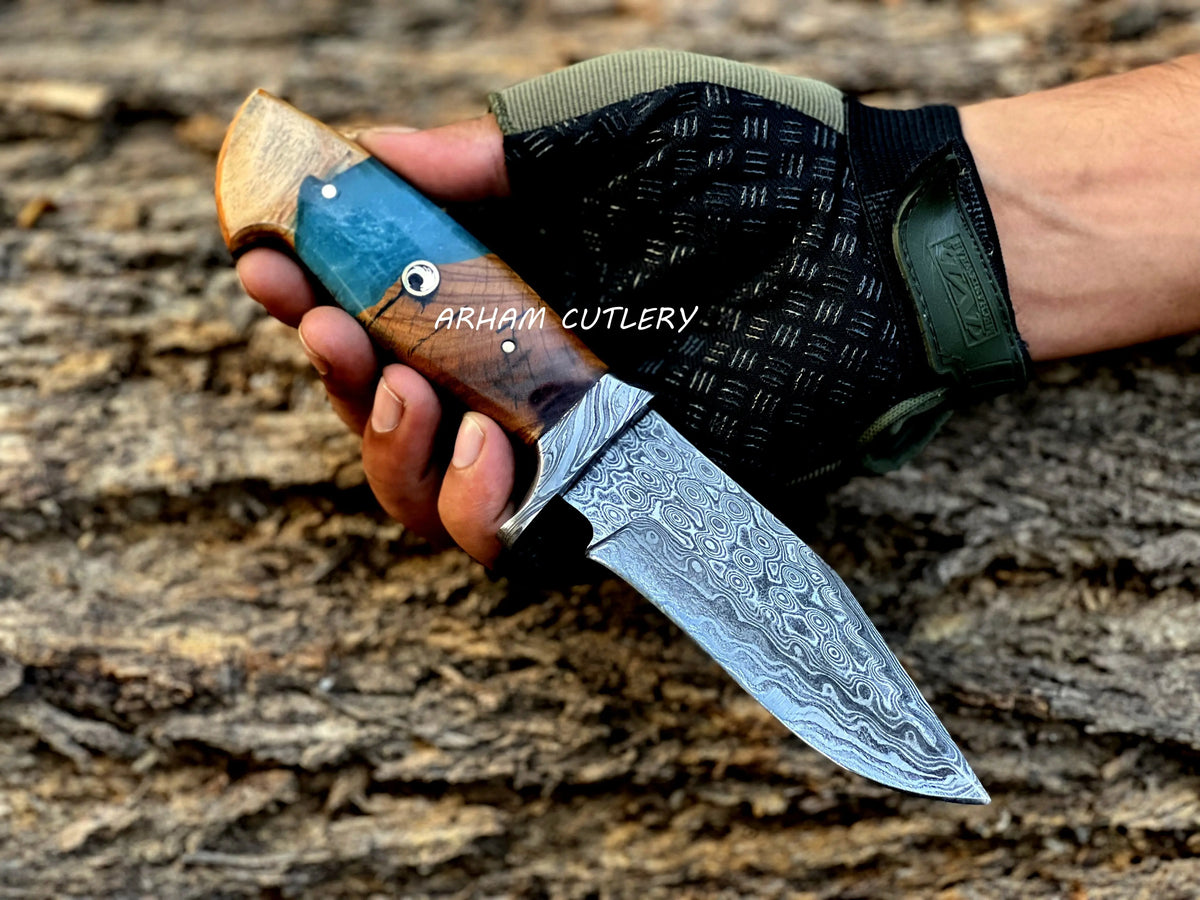 Hand-forged Damascus steel knife with epoxy and wood handle and custom leather sheath by Arham Cutlery, displayed on a wooden surface.