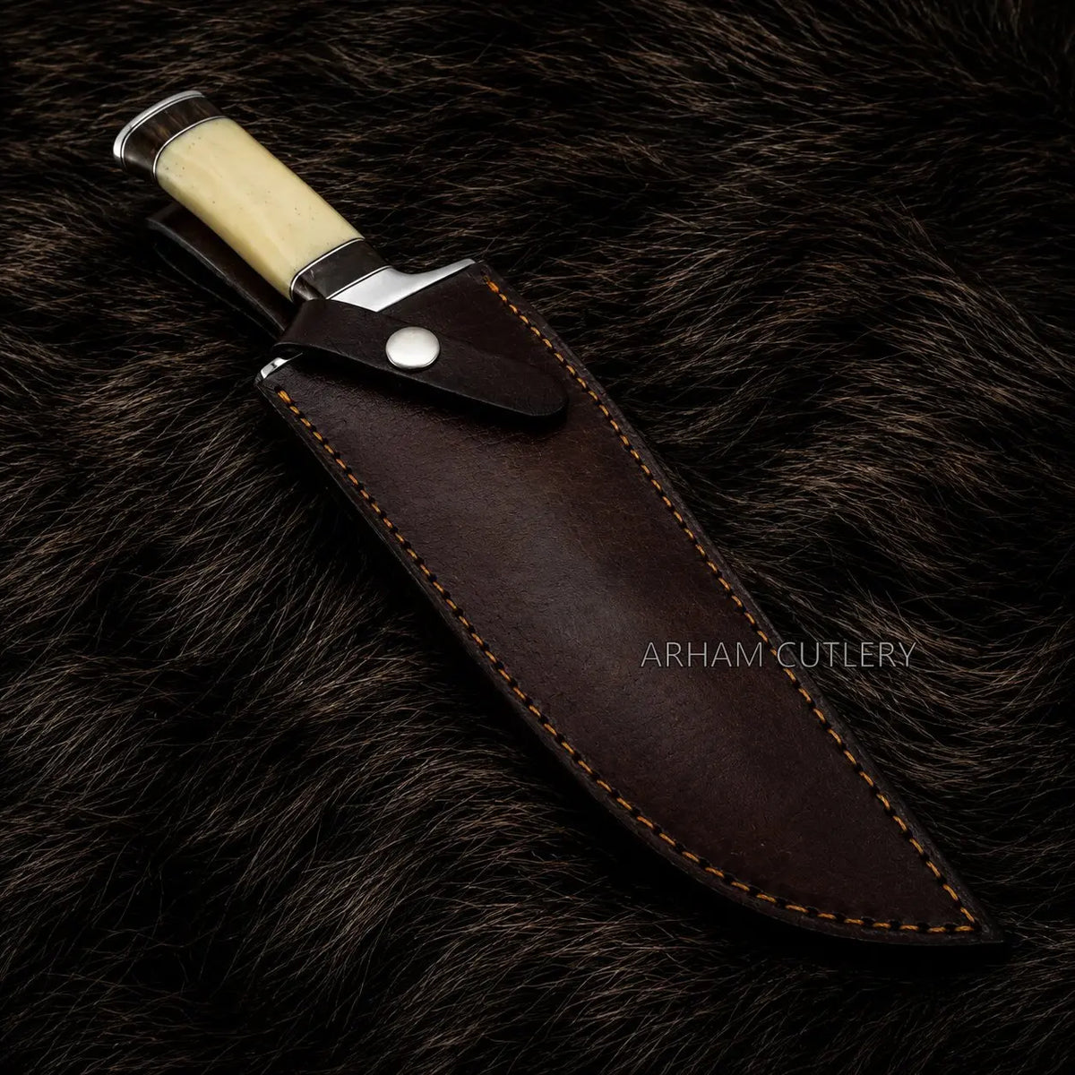 A fixed-blade knife with a cream-colored handle and silver bolster, shown partially sheathed in a dark brown leather sheath with contrast stitching, placed on a dark brown fur background. The brand name 'ARHAM CUTLERY' is watermarked on the sheath.