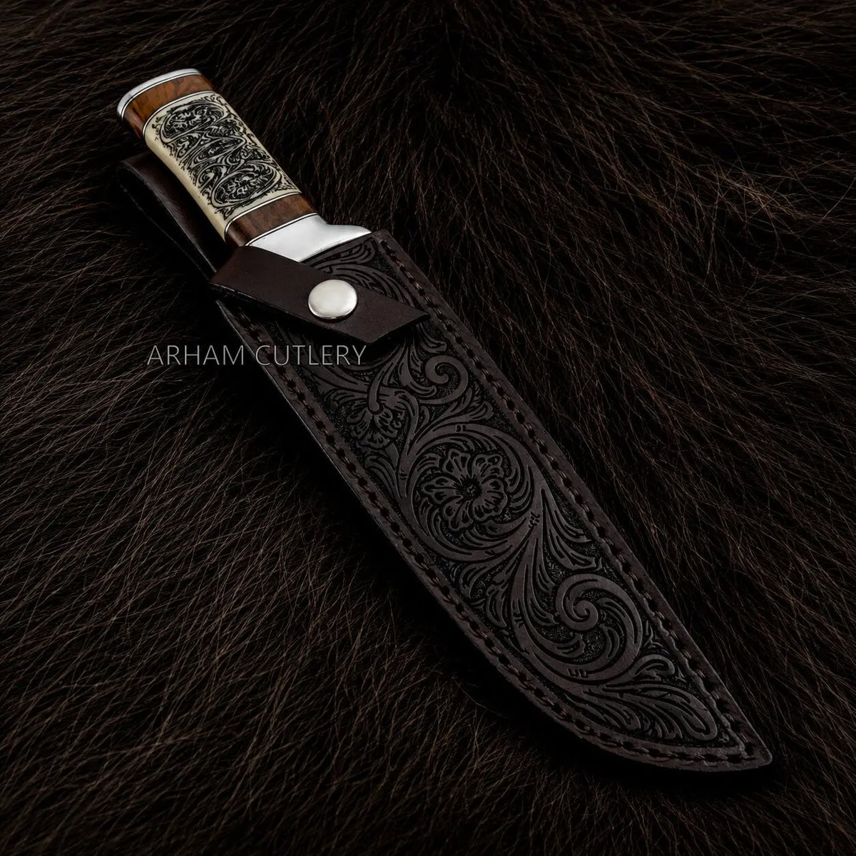 An ornate fixed-blade knife with a dark brown tooled leather sheath, displayed on a dark brown fur background. The knife features a silver-toned metal bolster, a silver engraved handle scale, and a wood-toned handle.