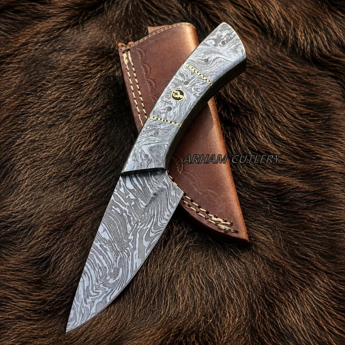 A Damascus steel fixed-blade knife with a wavy gray pattern blade, a brown leather sheath, and brass hardware, displayed diagonally on a brown fur background. A watermark for 'ARHAM CUTLERY' is visible across the center.