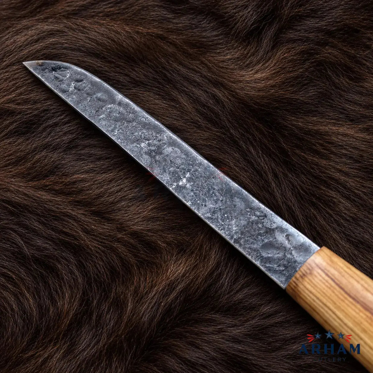 A close-up of a knife with a textured, hammered steel blade and a light brown wooden handle, laid diagonally on a dark brown fur background. The ARHAM brand logo is visible in the bottom right corner.