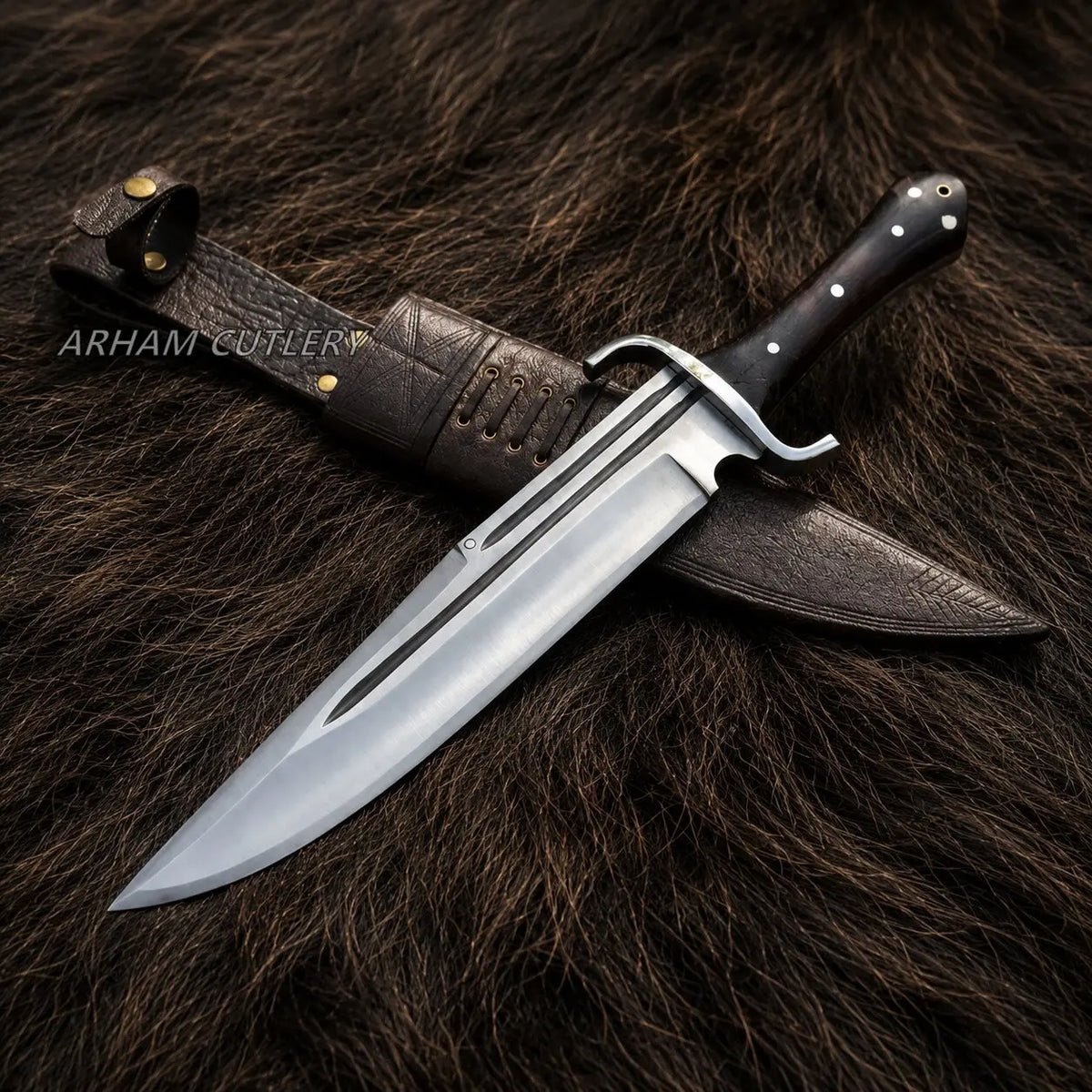 A fixed-blade knife with a polished silver blade, black handle, and brass guard, displayed diagonally with its dark brown leather sheath on a dark brown fur background. The brand name 'ARHAM COTLERY' is watermarked on the left.