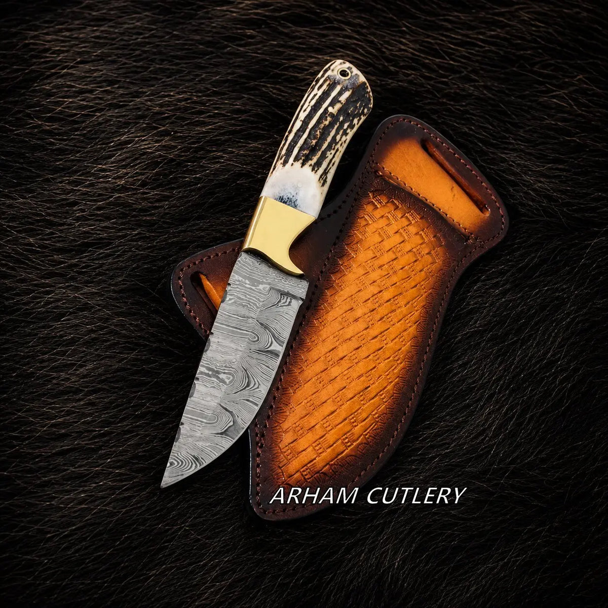 A fixed-blade knife with a patterned steel blade, a natural antler handle, and a brass bolster, shown resting in a brown leather sheath on a dark textured fur background. The text 'ARHAM CUTLERY' is visible at the bottom.