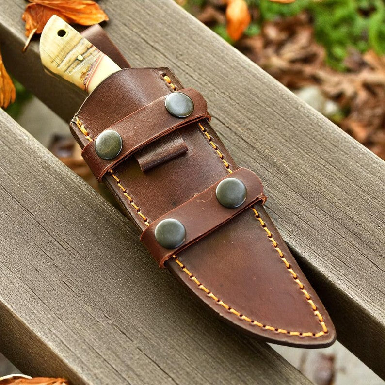 D2 steel knife with ram horn and camel bone handle in leather sheath on wooden bench.