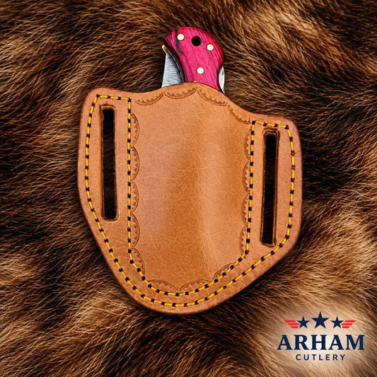 A tan leather knife sheath with decorative stitching and a pink handle partially inserted, photographed on brown fur. The Arham Cutlery logo appears in the bottom right corner.