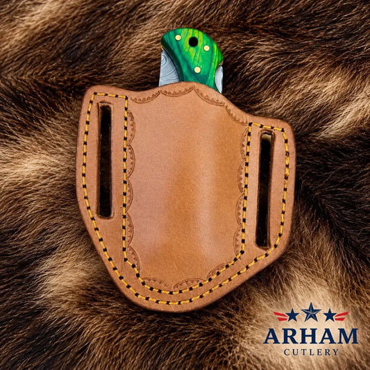 A tan leather knife sheath with decorative stitching and a green marbled knife handle partially inserted, photographed on a brown fur background. The Arham Cutlery logo appears in the bottom right corner.