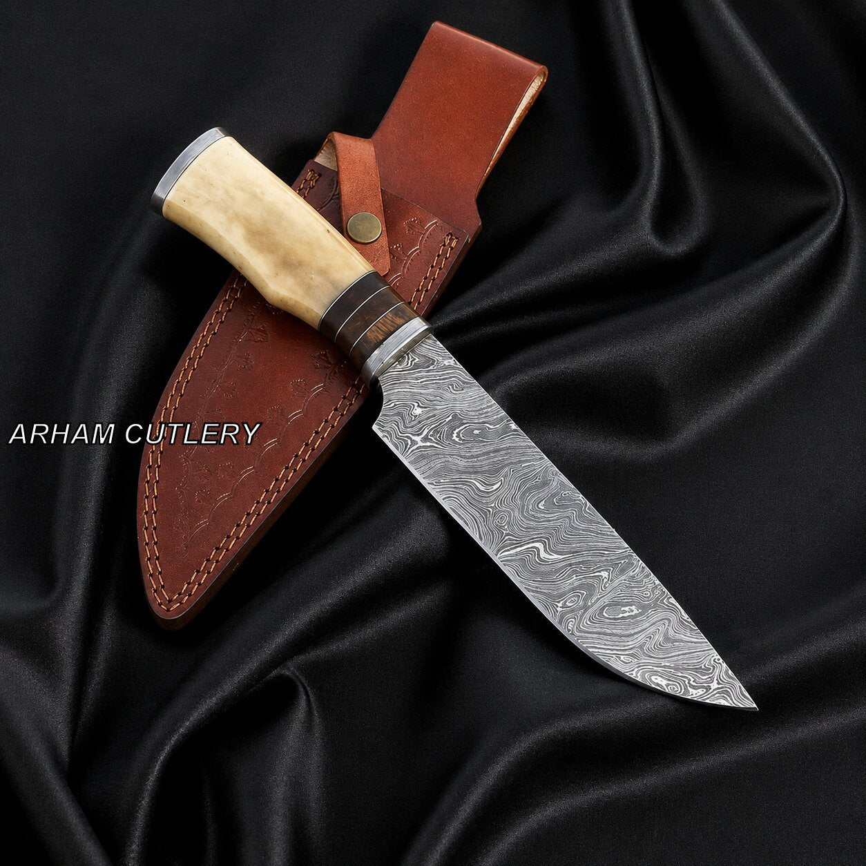 A Damascus steel fixed-blade knife with a light wood handle and a brown leather sheath, displayed on draped black satin fabric. The brand name 'ARHAM CUTLERY' is visible as a watermark on the left.