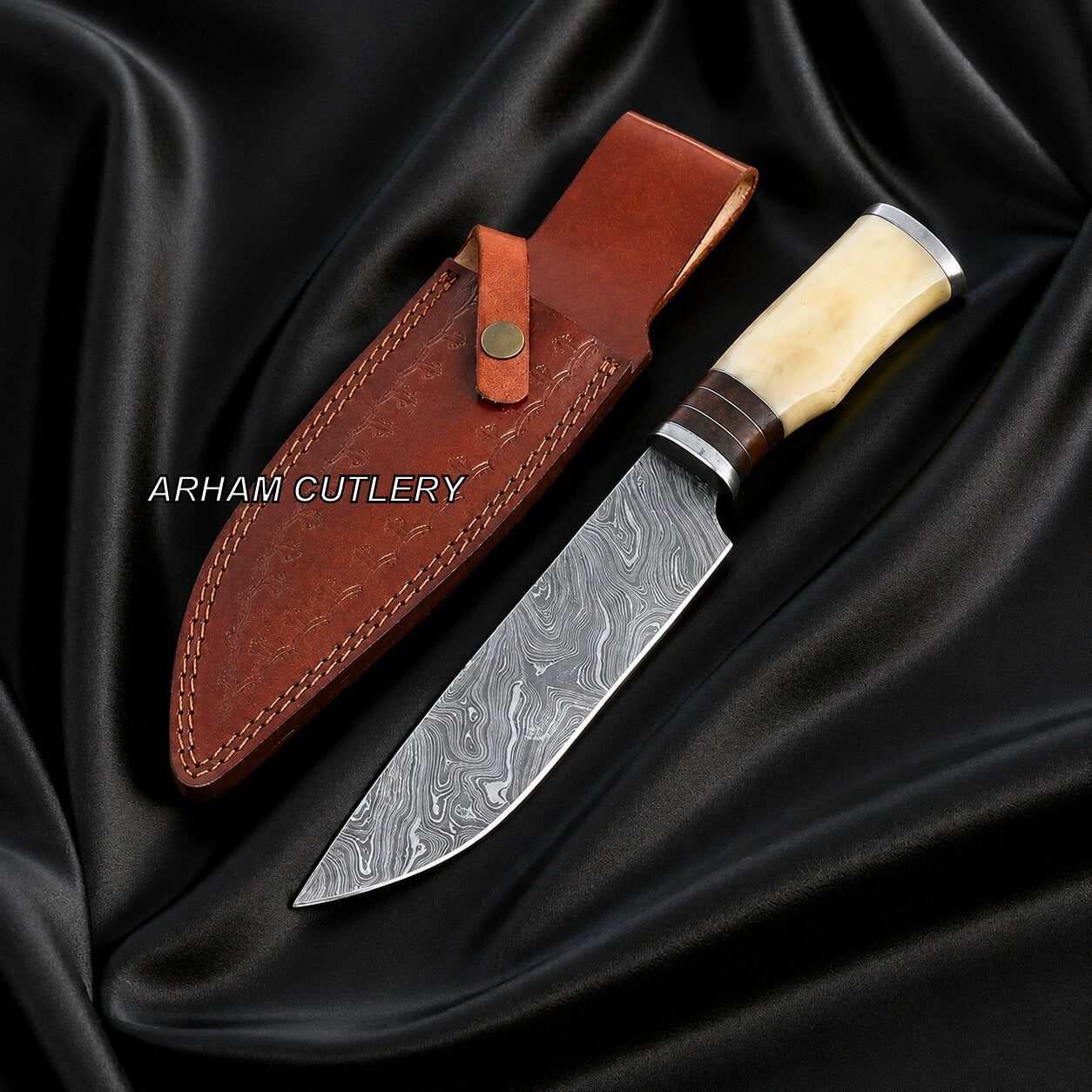 A fixed-blade knife with a patterned steel blade, cream bone handle, and silver bolster, displayed next to its brown leather sheath on a draped black satin fabric background. The brand name 'ARHAM CUTLERY' is printed in white text on the left.