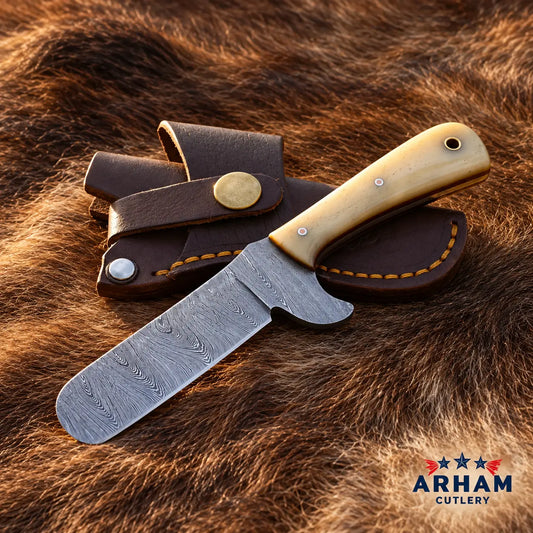 A fixed-blade knife with a patterned steel blade and a light tan bone handle, displayed next to its brown leather sheath on a brown fur background. The ARHAM CUTLERY logo is visible in the bottom right corner.
