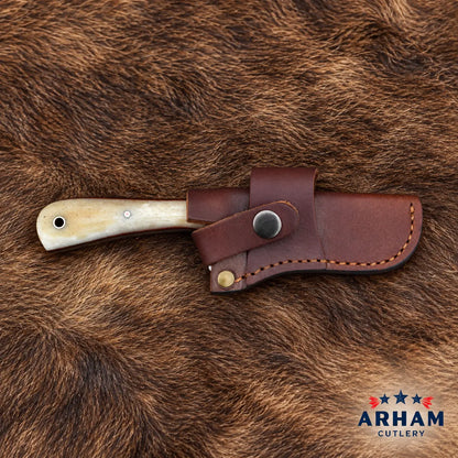 A folding knife with an ivory-colored bone handle and a dark brown leather sheath, displayed on a brown animal fur background. The ARHAM CUTLERY logo is visible in the bottom right corner.