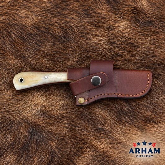 A folding knife with an ivory-colored bone handle and a dark brown leather sheath, displayed on a brown animal fur background. The ARHAM CUTLERY logo is visible in the bottom right corner.
