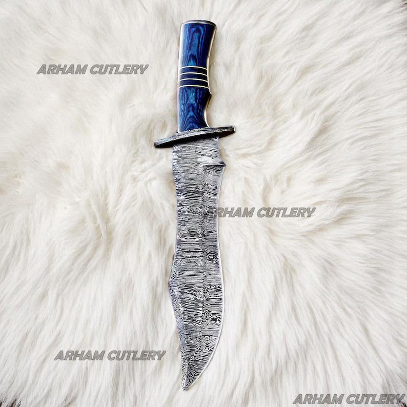 Hand-forged Damascus steel Bowie knife with blue Pakka Wood handle and custom leather sheath displayed on tree bark outdoors