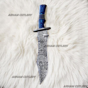 Hand-forged Damascus steel Bowie knife with blue Pakka Wood handle and custom leather sheath displayed on tree bark outdoors