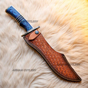 Hand-forged Damascus steel Bowie knife with blue Pakka Wood handle and custom leather sheath displayed on tree bark outdoors