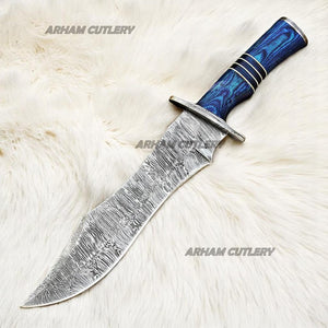 Hand-forged Damascus steel Bowie knife with blue Pakka Wood handle and custom leather sheath displayed on tree bark outdoors