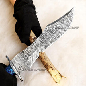 Hand-forged Damascus steel Bowie knife with blue Pakka Wood handle and custom leather sheath displayed on tree bark outdoors