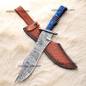 Hand-forged Damascus steel Bowie knife with blue Pakka Wood handle and custom leather sheath displayed on tree bark outdoors
