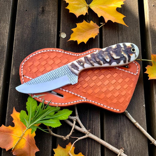 Frontier Fang ranch knife with sheep horn handle and crosshatched blade, displayed on a leather sheath and rustic wood background with fall leaves.