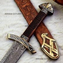 Cargar imagen en el visor de la galería, Hand-forged Viking Damascus steel sword with engraved wooden scabbard and ornate brass fittings displayed on a dark wooden background
