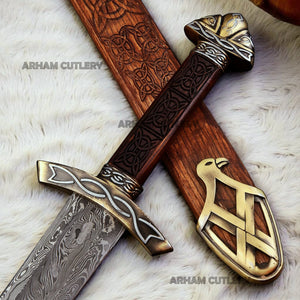 Hand-forged Viking Damascus steel sword with engraved wooden scabbard and ornate brass fittings displayed on a dark wooden background