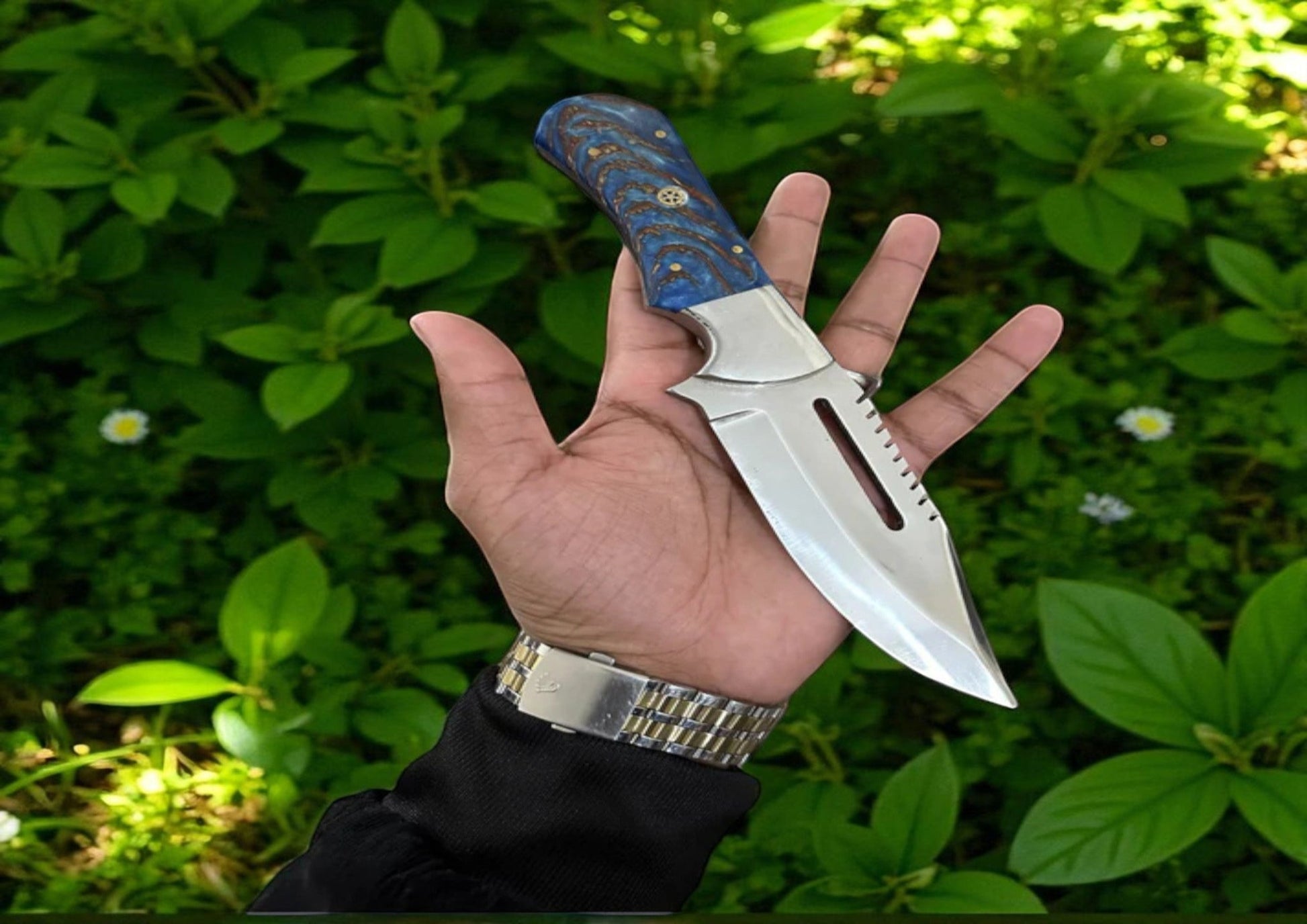 The Frostfang knife by Arham Cutlery featuring a blue pine cone resin handle and a D2 steel blade, displayed in hand against a green foliage background.

