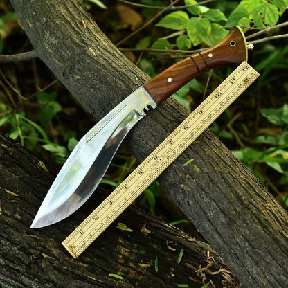 A large fixed-blade knife with a polished silver blade and a brown wooden handle, shown resting on a tree branch outdoors next to a wooden ruler for scale.