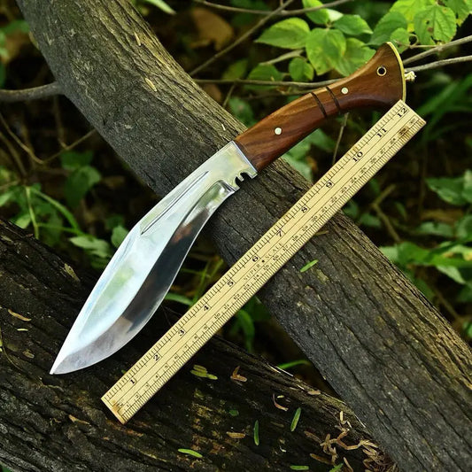 A large fixed-blade knife with a polished silver blade and a brown wooden handle, shown resting on a tree branch outdoors next to a wooden ruler for scale.