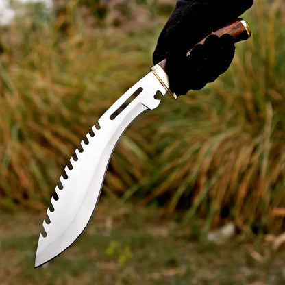 A curved silver machete-style knife with serrated edges, held by a hand wearing a black glove against a blurred outdoor background of dry grass.