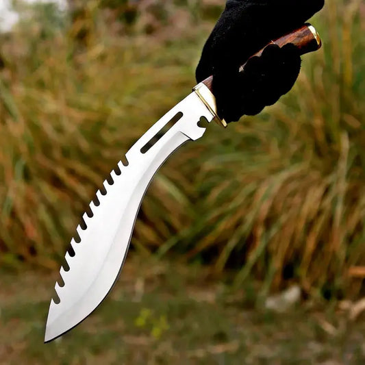 A curved silver machete-style knife with serrated edges, held by a hand wearing a black glove against a blurred outdoor background of dry grass.