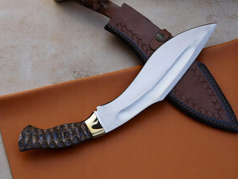 A kukri-style knife with a polished silver blade, textured dark brown handle, and gold-colored bolster, displayed diagonally on a brown leather sheath over a tan surface.