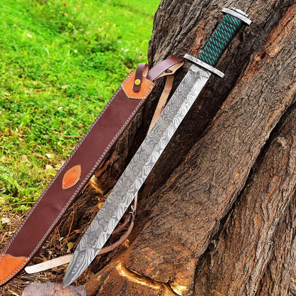A patterned steel sword with a green wrapped handle and brown leather sheath, leaning against a tree trunk outdoors on grass.
