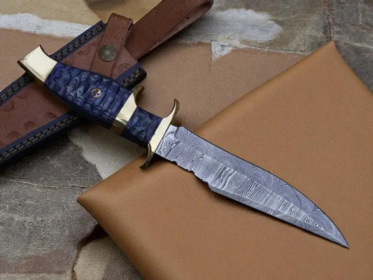A fixed-blade knife with a patterned steel blade, textured blue handle scales, and brass guard, displayed diagonally on a brown leather sheath and tan surface.