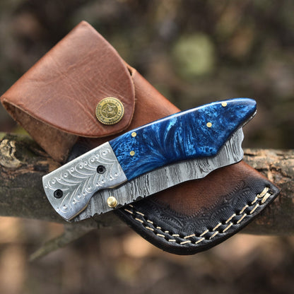 A folding pocket knife with a blue marbled handle and patterned silver blade, resting on a brown leather sheath with a blurred outdoor background.