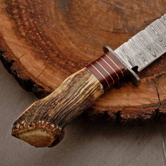 A close-up of a handcrafted knife with a patterned Damascus steel blade, a reddish-brown wrapped guard, and a natural antler handle, resting on a rustic wood slice background.